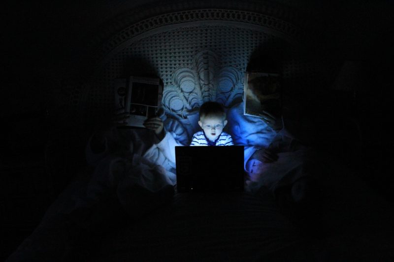 cara mempercepat koneksi internet, anak sedang bermain laptop di kamar orang tuanya dengan lampu yang mati dan gelap