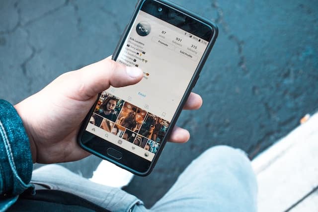 Seorang yang menggunakan baju biru dengan tangan yang sedang memegang handphone di jalan dengan layar ponsel menyala membuka aplikasi instagram