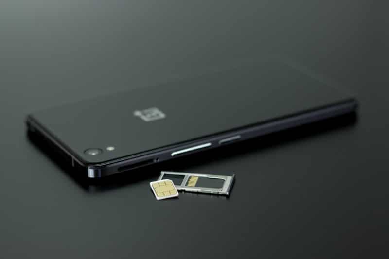 handphone android berwarna hitam tergeletak di meja dengan posisi telungkup layar menghadap bawah dan slot kartu sim yang dikeluarkan bersama sim card di atasnya, background hitam